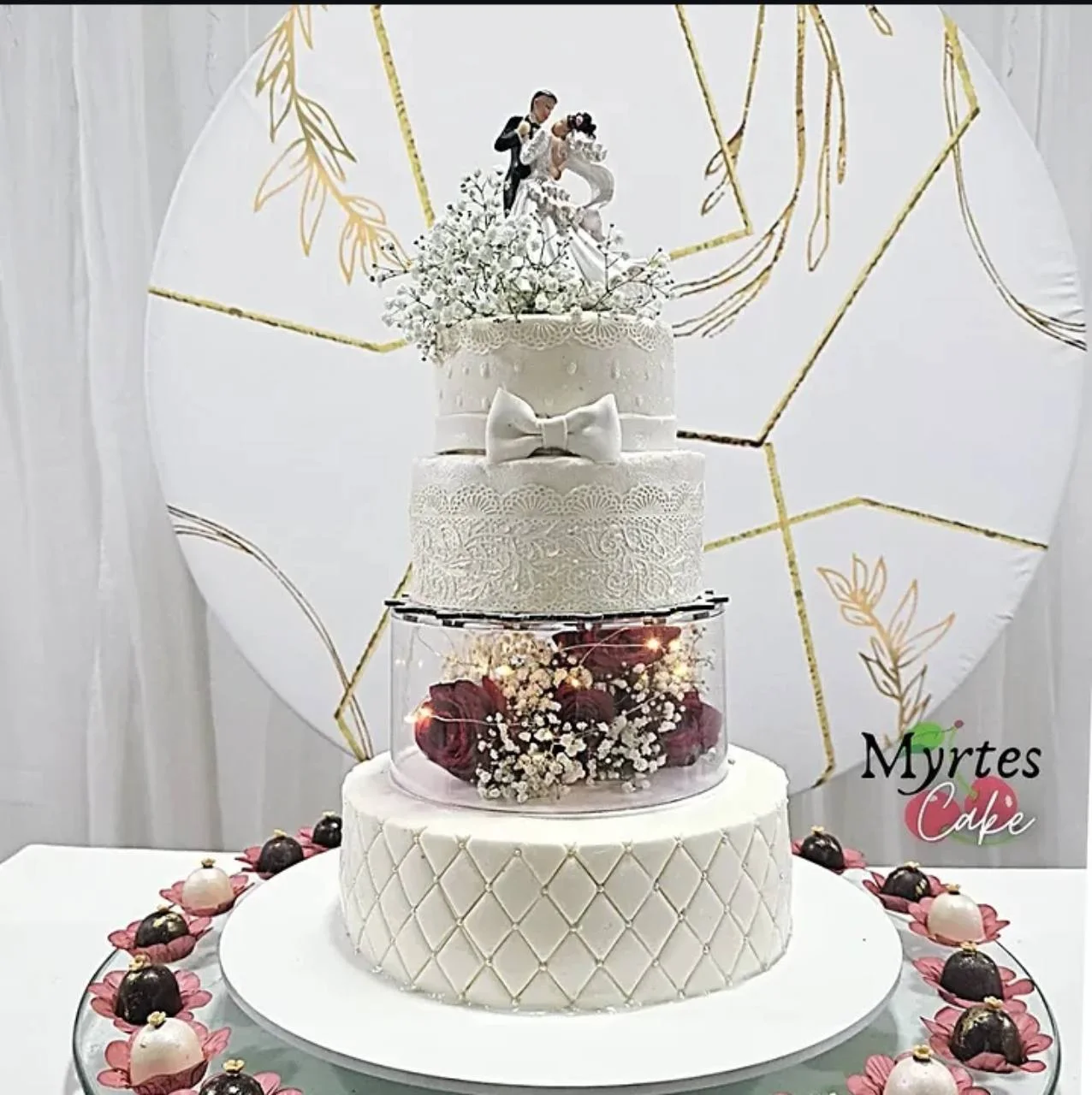 Bolo branco de andares com laço, detalhes de renda, flores e topo com casal, sobre uma mesa decorada.